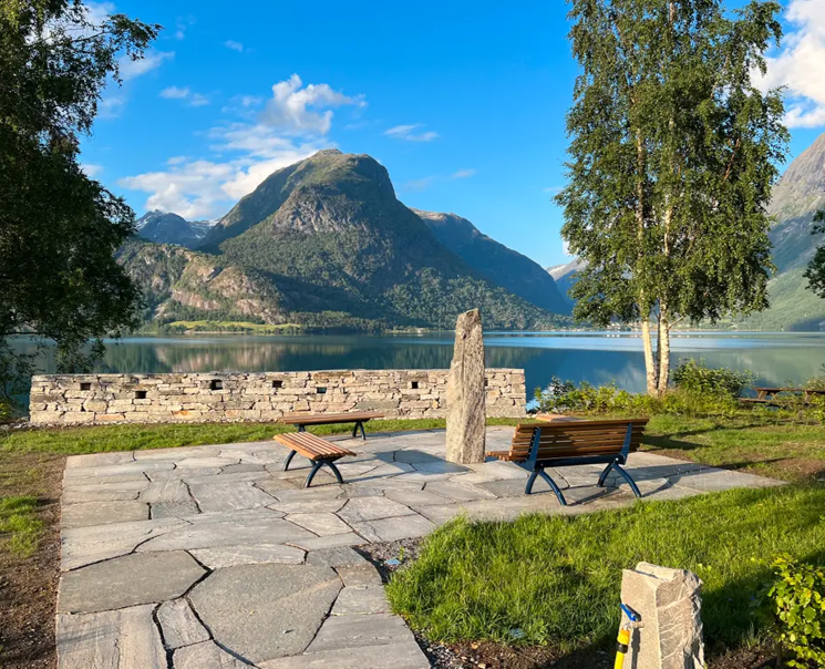 Bilde viser den namna Minnelunden i Oppstryn som er fint opparbeidd med skiferheller med grøne plen rundt til askeurner. Der er ein store høge bautastein midt på skiferplassen til å ha namn til dei avdøde på. Der er også trebenkar og ein mur ned mot vatnet til å ha gravlykter i.  Vi ser også Oppstrynsvatnet og dei flotte høge fjella i bakgrunnen ein solrik sommarsdag.